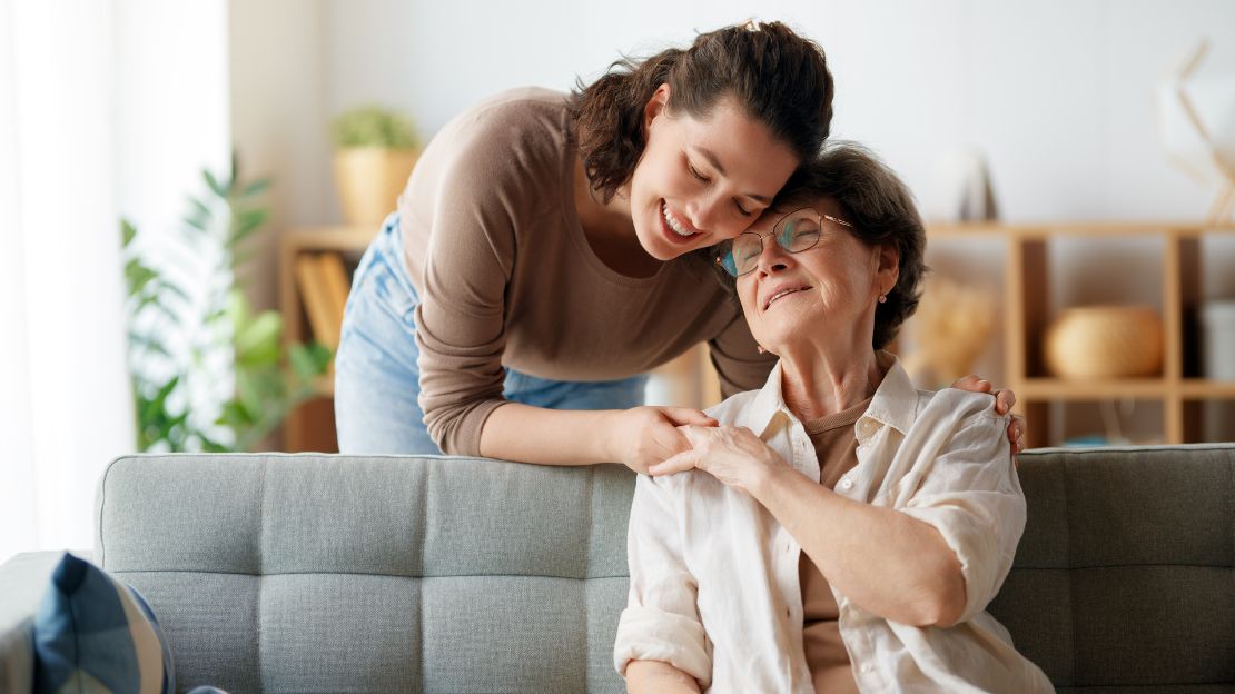 Das Bild zeigt eine junge Betreuerin, die eine Seniorin in deren Wohnung in dem Arm nimmt.