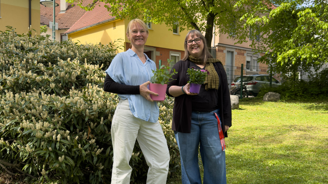 Tina Wenk links und Johanna Greef stehen im Freien vor einem Wohngebäude und halten jeweils einen Blumentopf mit Pflanzen in den Händen. Beide lächeln in die Kamera, im Hintergrund sind Sträucher und ein Baum zu sehen.
