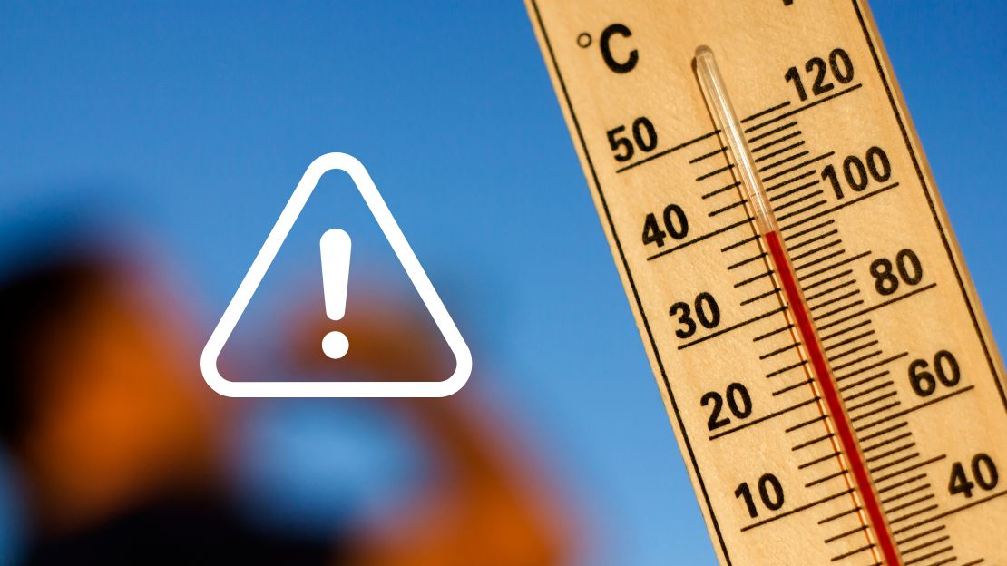 Nahaufnahme eines Thermometers mit hoher Temperaturanzeige vor blauem Himmel. Ein weißes Warnsymbol mit Ausrufezeichen weist auf die gesundheitlichen Risiken großer Hitze hin.