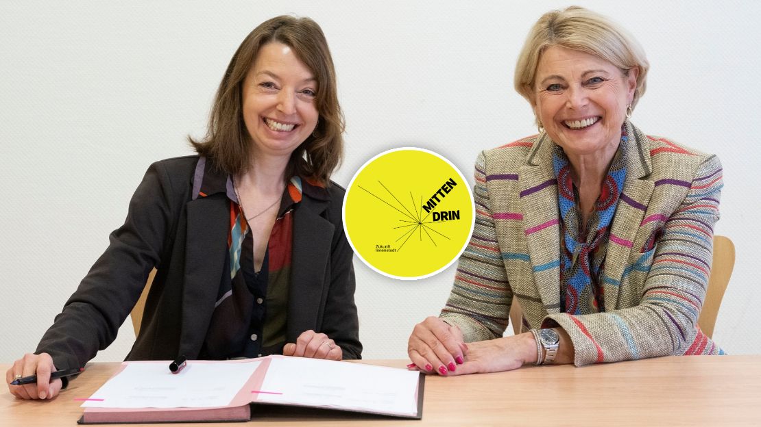Nadine Bernecker und Agnes Christner sitzen am Tisch vor den unterzeichneten Vertragsunterlagen. Beide Frauen lächeln frlöhlich in die Kamera. In der Linken Ecke des Bildes ist das Logo "Mittendrin" für das Sofortprogramm Zukunft Innenstadt abgebildet.