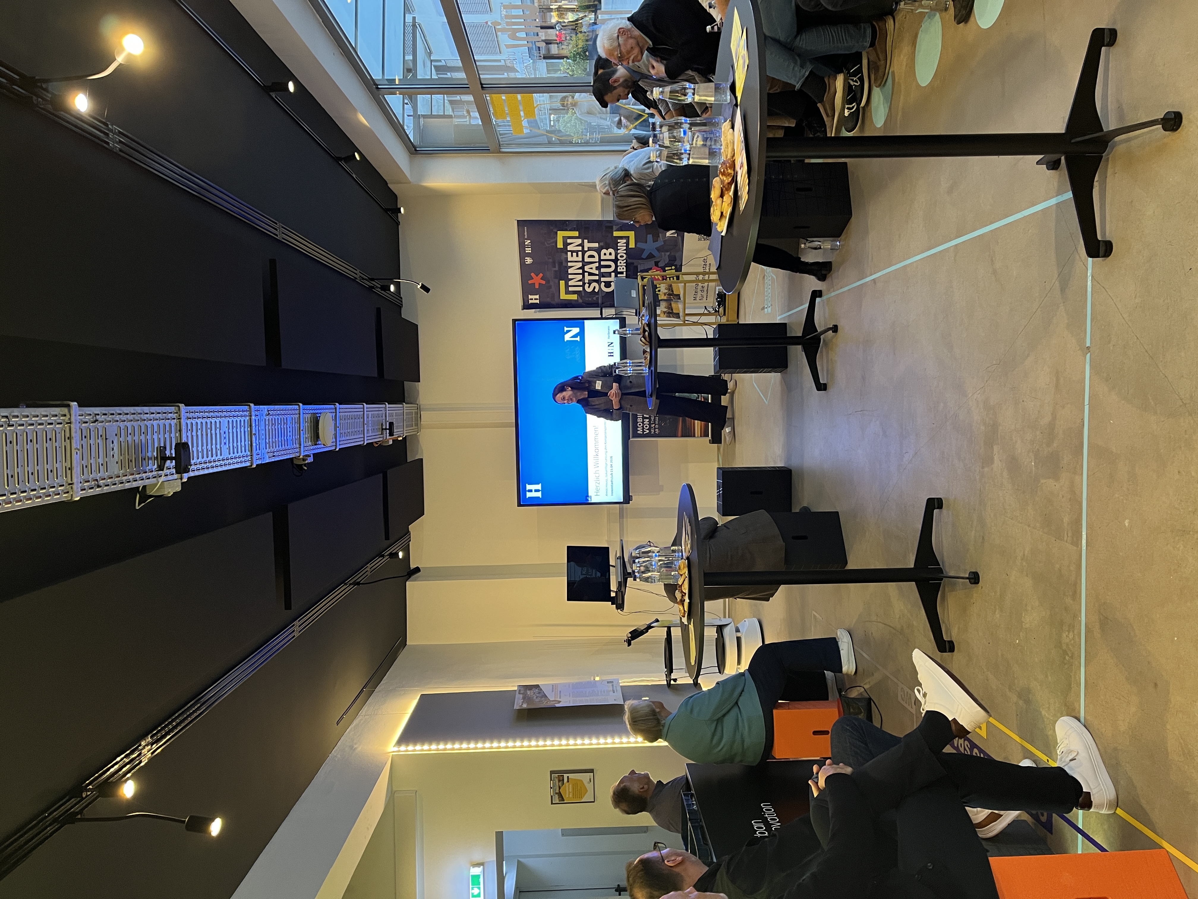 Vortragssituation in einem modernen Veranstaltungsraum: Nadine Izquierdo präsentiert vor Publikum das geplante Vorgehen für das Zukunftskonzept Innenstadt. Die Teilnehmenden sitzen an Stehtischen, im Hintergrund sind Präsentationsfolie und ein Banner zum Innenstadt Club Heilbronn zu sehen.